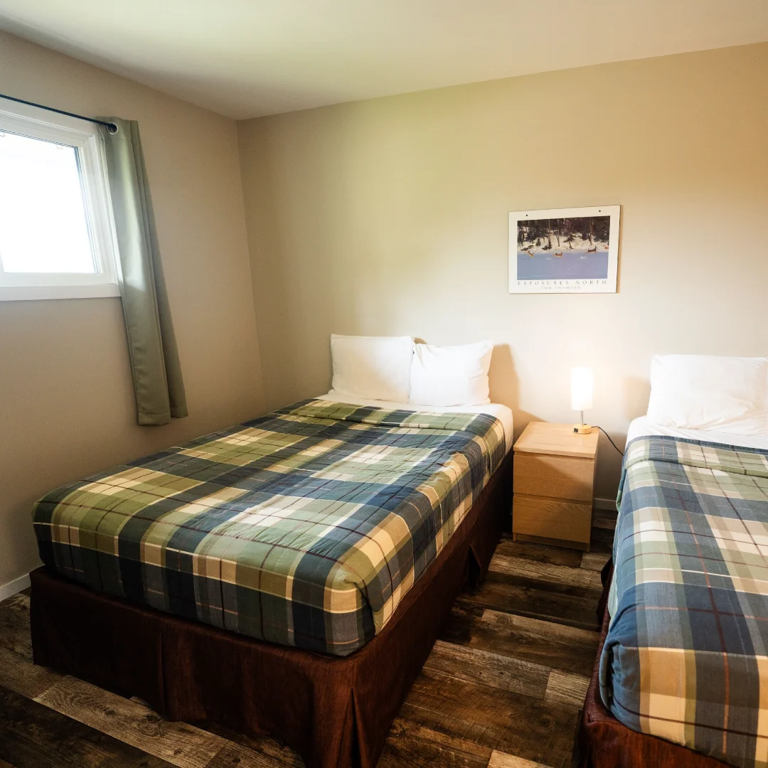 Suite bedroom with two beds, plaid blankets, white pillows, a nightstand, wall art, and warm lighting.
