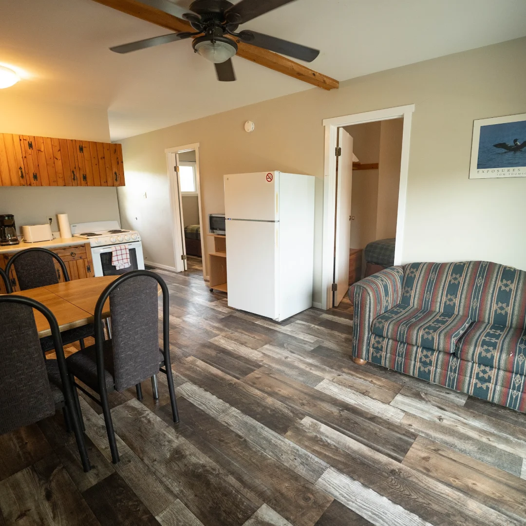 Open suite living area with a small kitchen, dining table, refrigerator, patterned sofa, wood-look flooring, and two bedroom doors.
