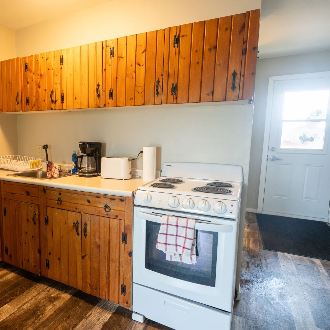 Suite kitchen with wood cabinets, white stove, sink, coffee maker, toaster, and a door leading outside.
