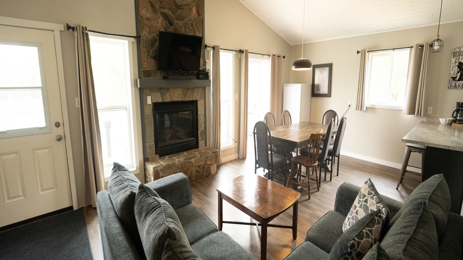 Interior of the Perch Bay Dam View Cabin with grey sofas, a wooden dining table, stone fireplace, and bright windows.
