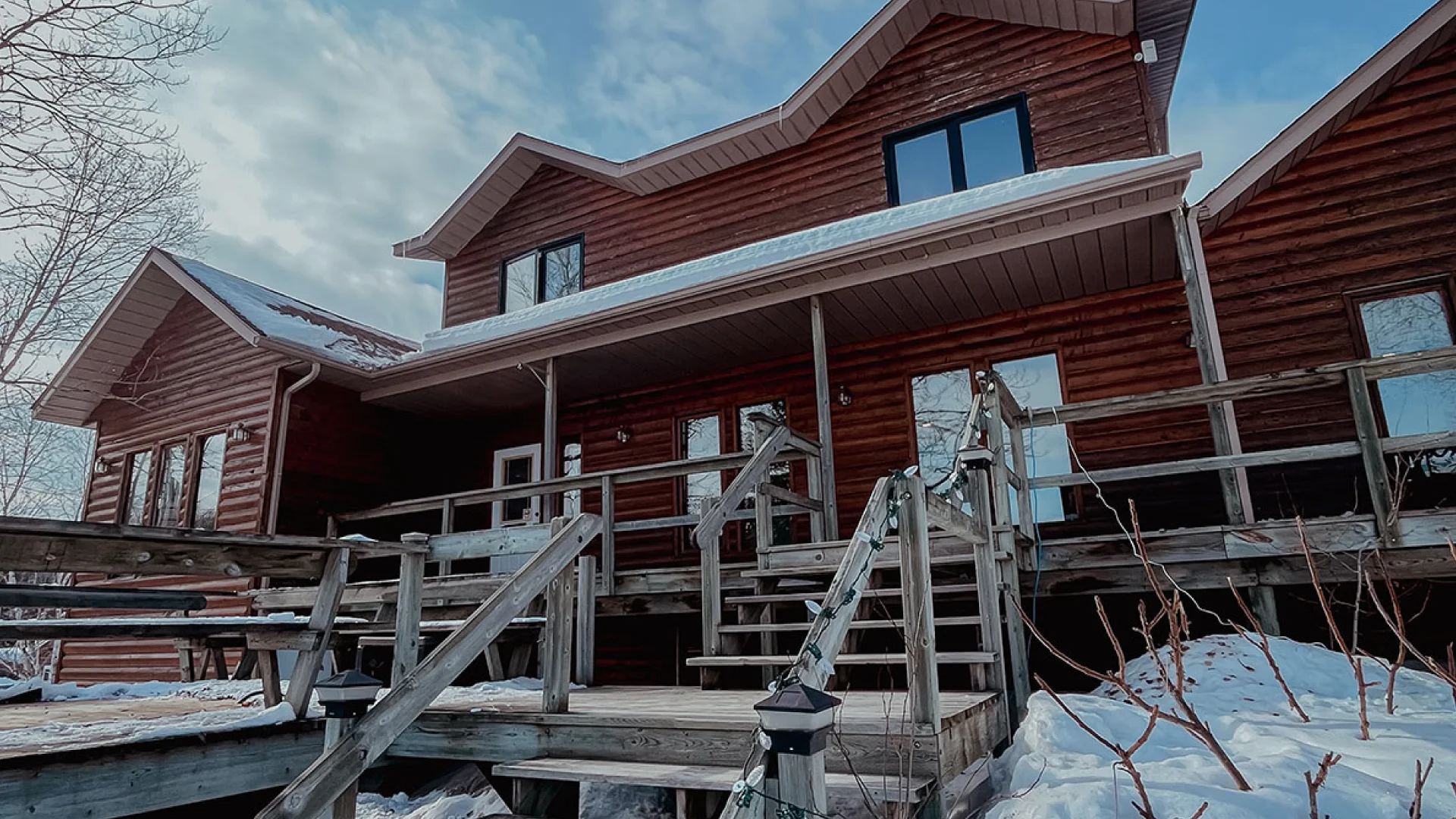 Winter view of the Perch Bay lodge suite exterior with log siding, wooden stairs, snow, and a blue sky overhead.