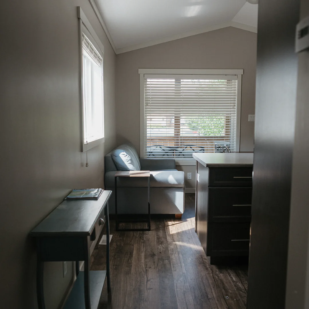 Small living area inside the Perch Bay Harbour Cabin with a grey sofa, side table, window blinds, and kitchen counter nearby.