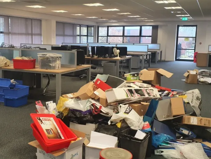 Office space with desks and computers in the background and a large cluttered pile of various boxes, folders, and packaging materials on the floor in the foreground.