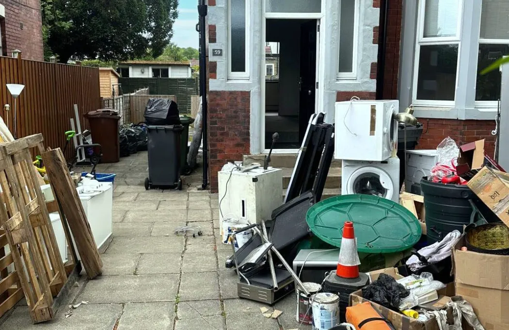 Backyard with various discarded household items including a washing machine, traffic cone, wooden pallets, and trash bins near a red brick house.