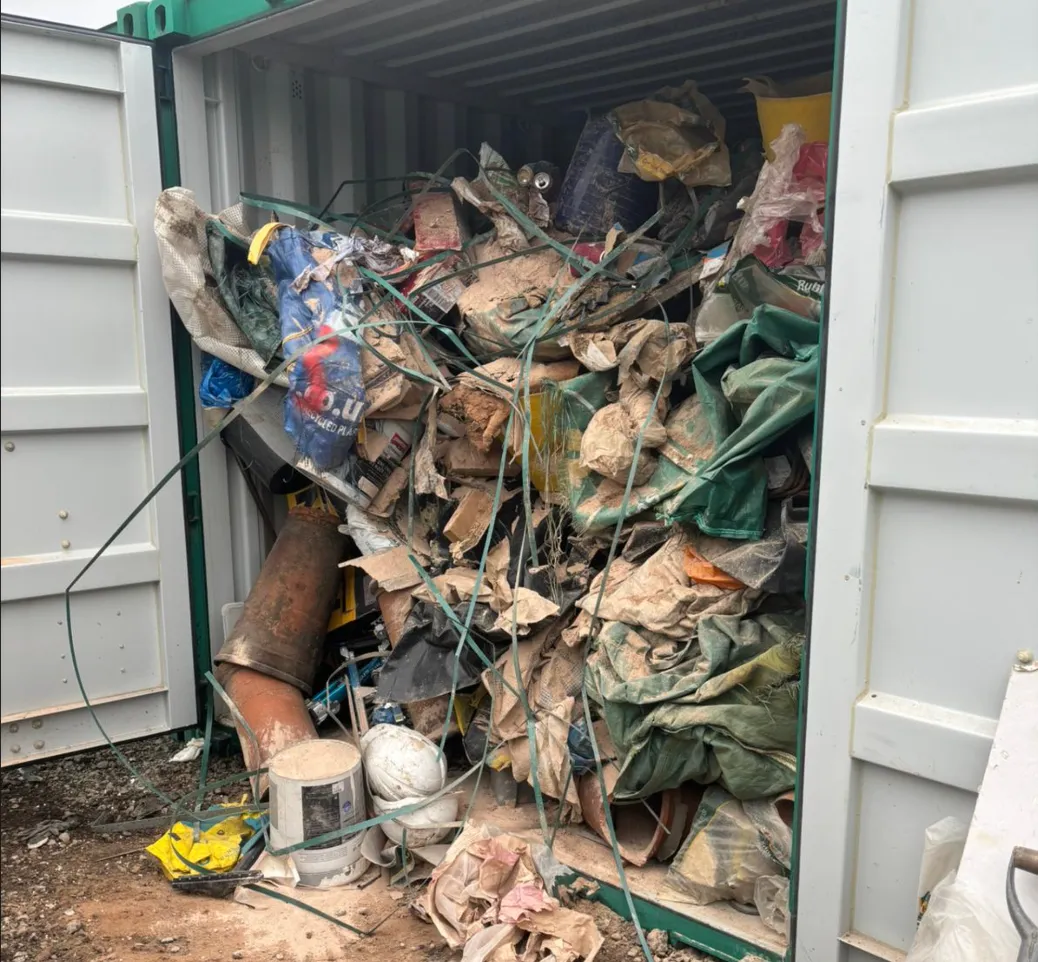 Open container filled with tightly packed construction debris, dirt, broken pipes, buckets, sacks, and plastic wrapping.