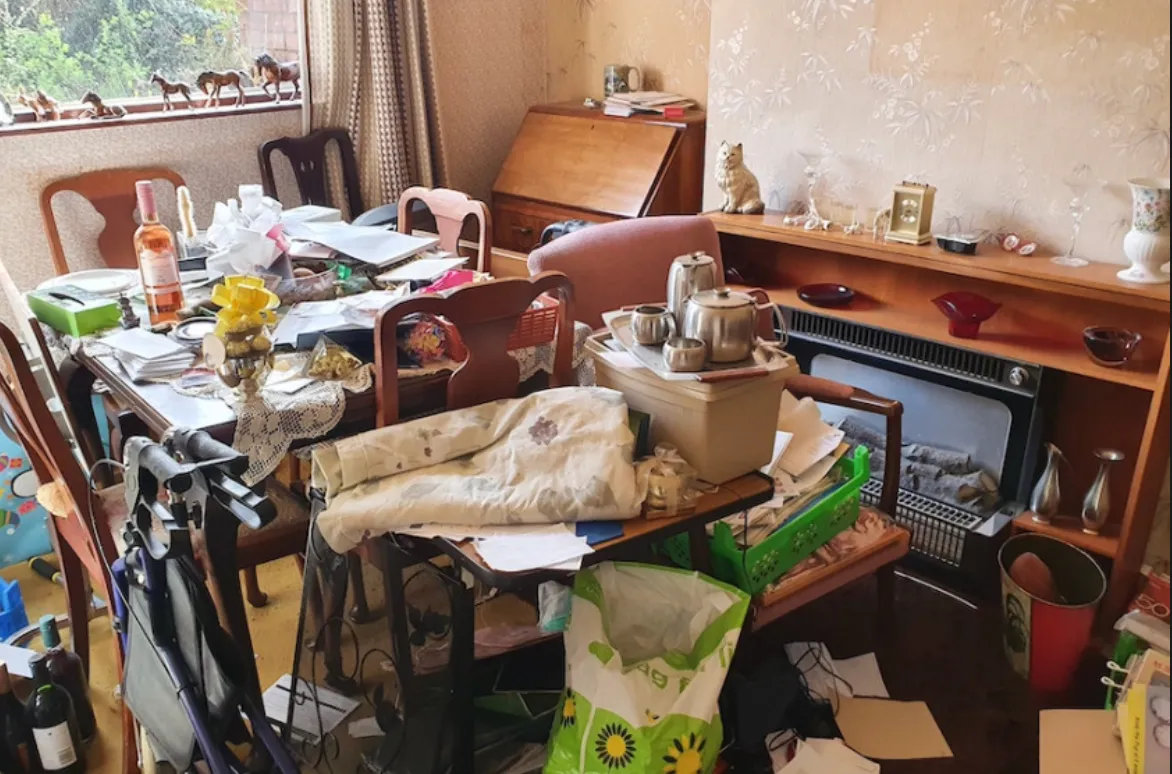 Cluttered room with a dining table covered in papers, bottles, and utensils, a walker, and various items scattered around.