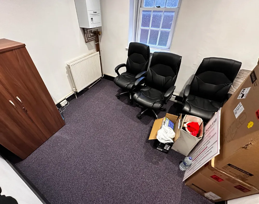 Small office corner with three black swivel chairs, wooden cabinet, cardboard boxes, and a water bottle on a purple carpeted floor under a window.