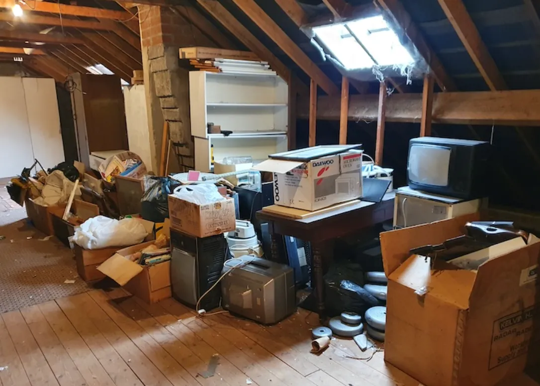Cluttered attic with wooden floor and exposed beams filled with boxes, vintage electronics including old TVs, and miscellaneous items.