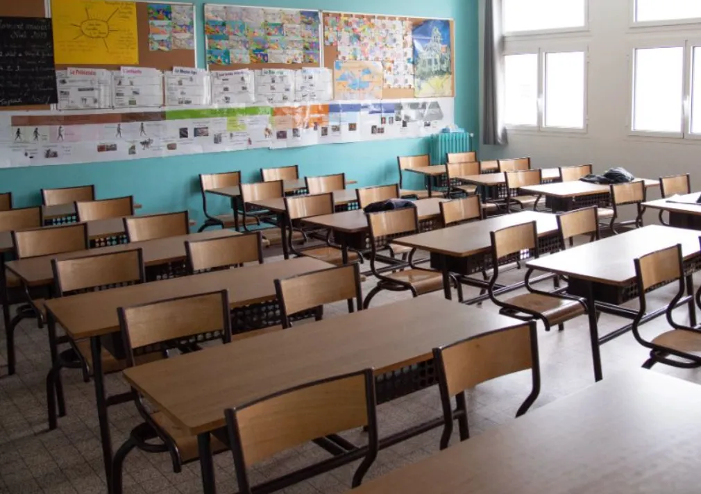 Empty classroom with rows of wooden desks and chairs, a bulletin board with colorful posters, and large windows letting in daylight.