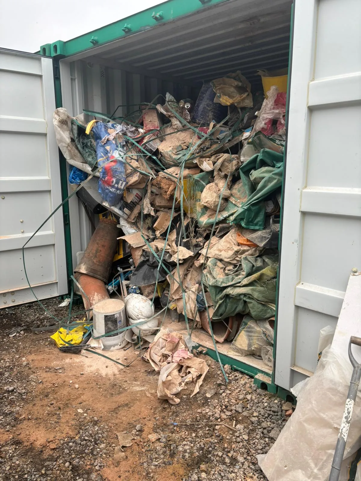 Open storage container filled with various construction debris, bags, pipes, and tangled green straps spilling onto a dirt and gravel ground.
