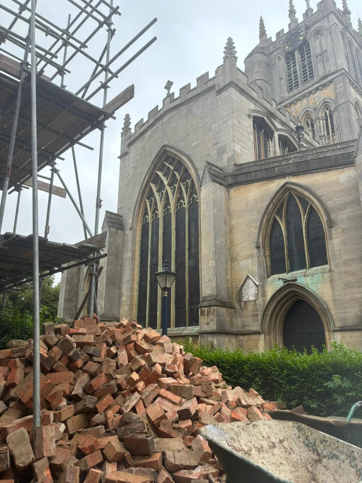 Pile of red bricks and scaffolding in front of a stone Gothic-style church with arched windows and a clock tower.