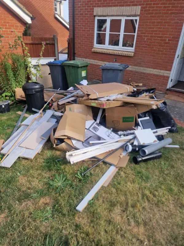 Pile of construction debris including cardboard, pipes, and wooden planks on grass near a brick house with multiple trash bins in the background.