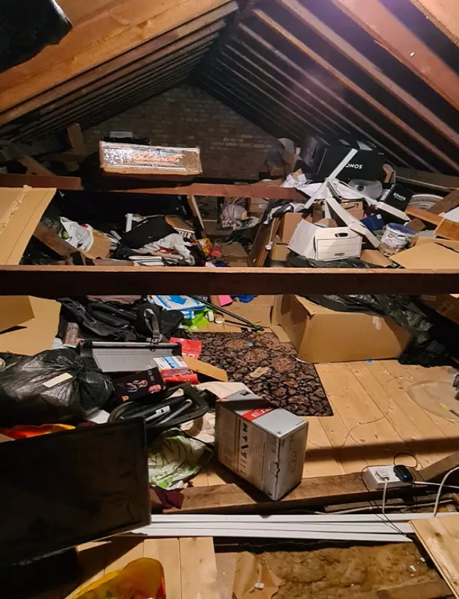 Cluttered attic with exposed wooden beams filled with various boxes, bags, and miscellaneous stored items on a wooden floor.