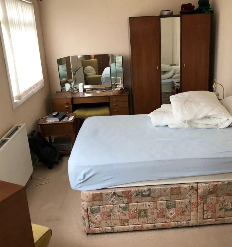 Cozy bedroom with a bed covered in light blue sheets and white bedding, wooden wardrobe with mirror, dressing table with three-panel mirror, and side table with remote controls.