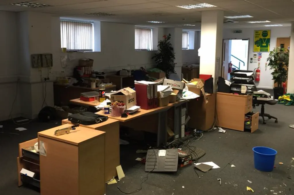 Cluttered office space with desks covered in scattered papers, boxes, and electronic cables, with some drawers open and a blue trash bin on the floor.