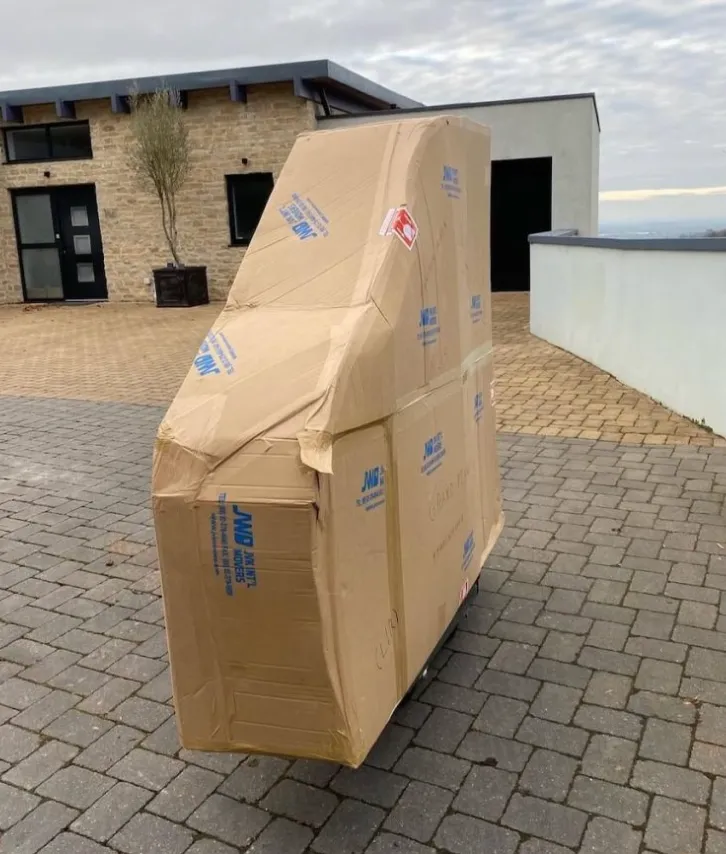 Large cardboard box with angled top standing on a paved driveway outside a modern house.