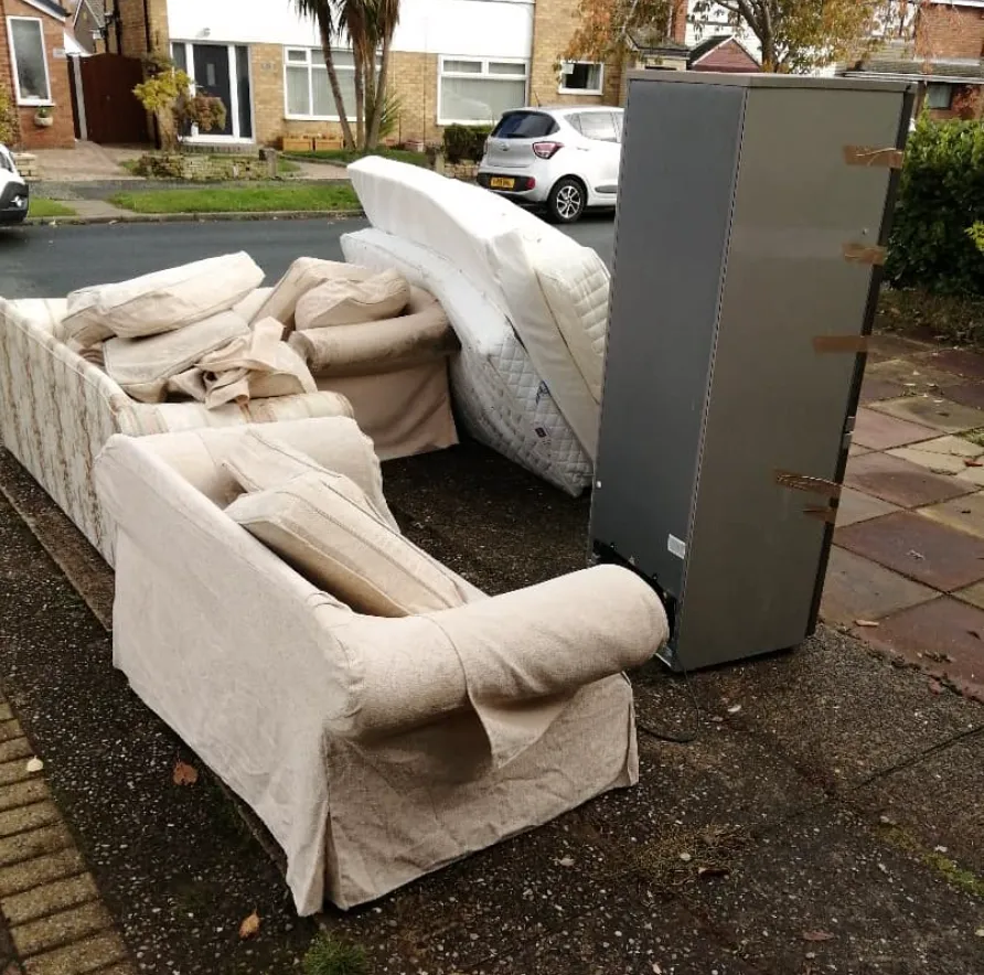 Discarded beige sofas, cushions, mattresses, and a large gray appliance on a residential sidewalk.
