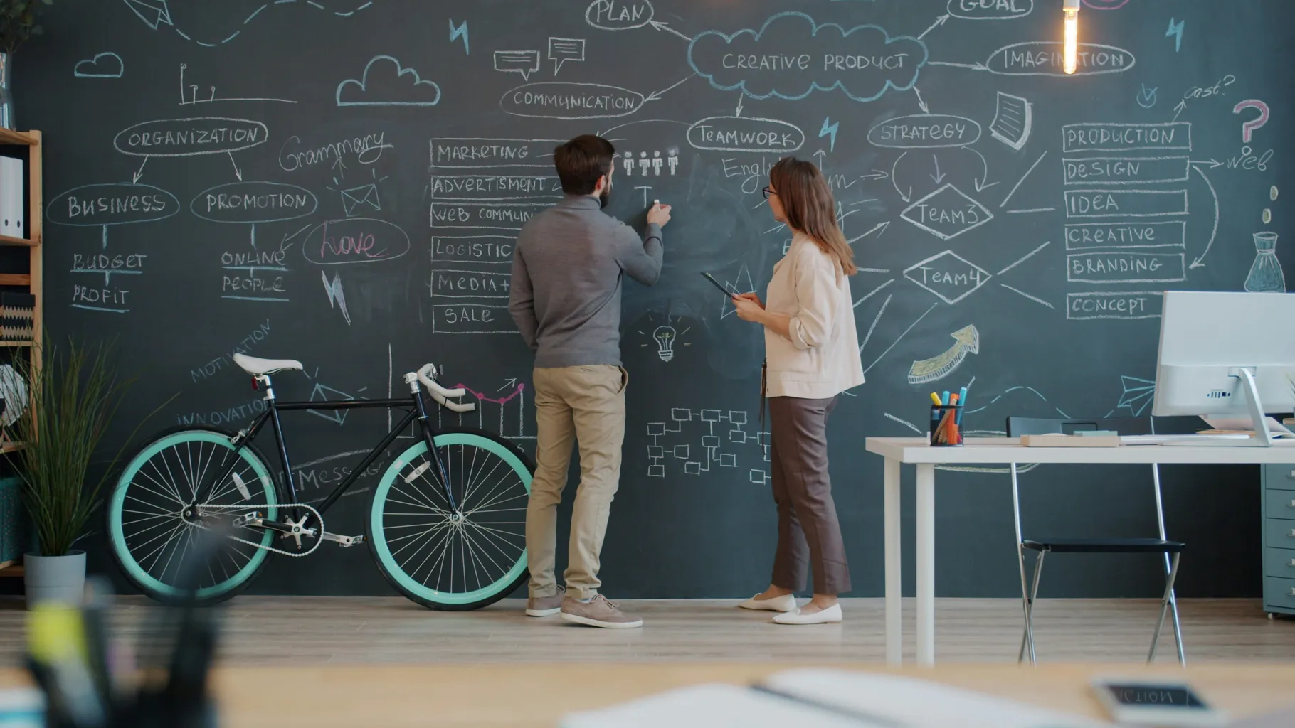 Two people collaborating and writing on a large chalkboard with business and marketing diagrams in a modern office.