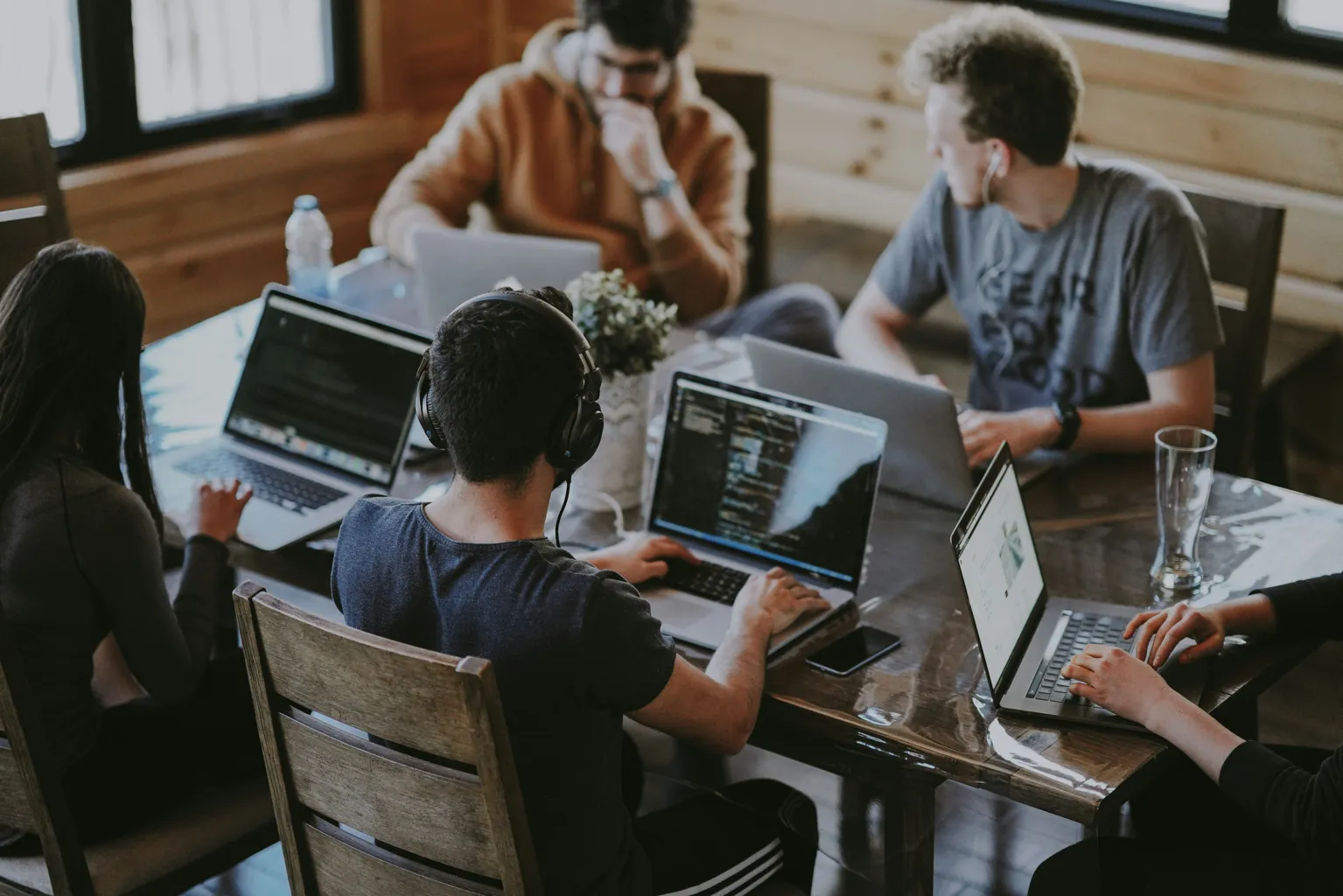 Group of five young adults working on laptops around a wooden table in a cozy room.