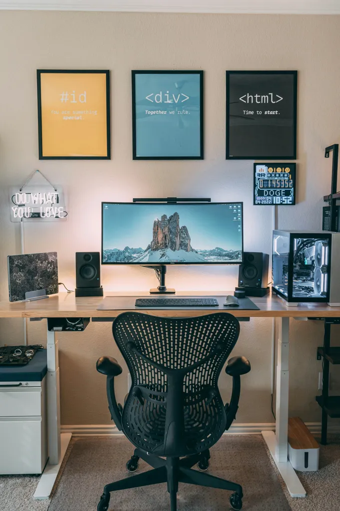 Modern computer workspace with an ultra-wide monitor displaying a mountain landscape, a black ergonomic office chair, framed coding-themed posters on the wall, speakers, and a transparent PC case on a standing desk.
