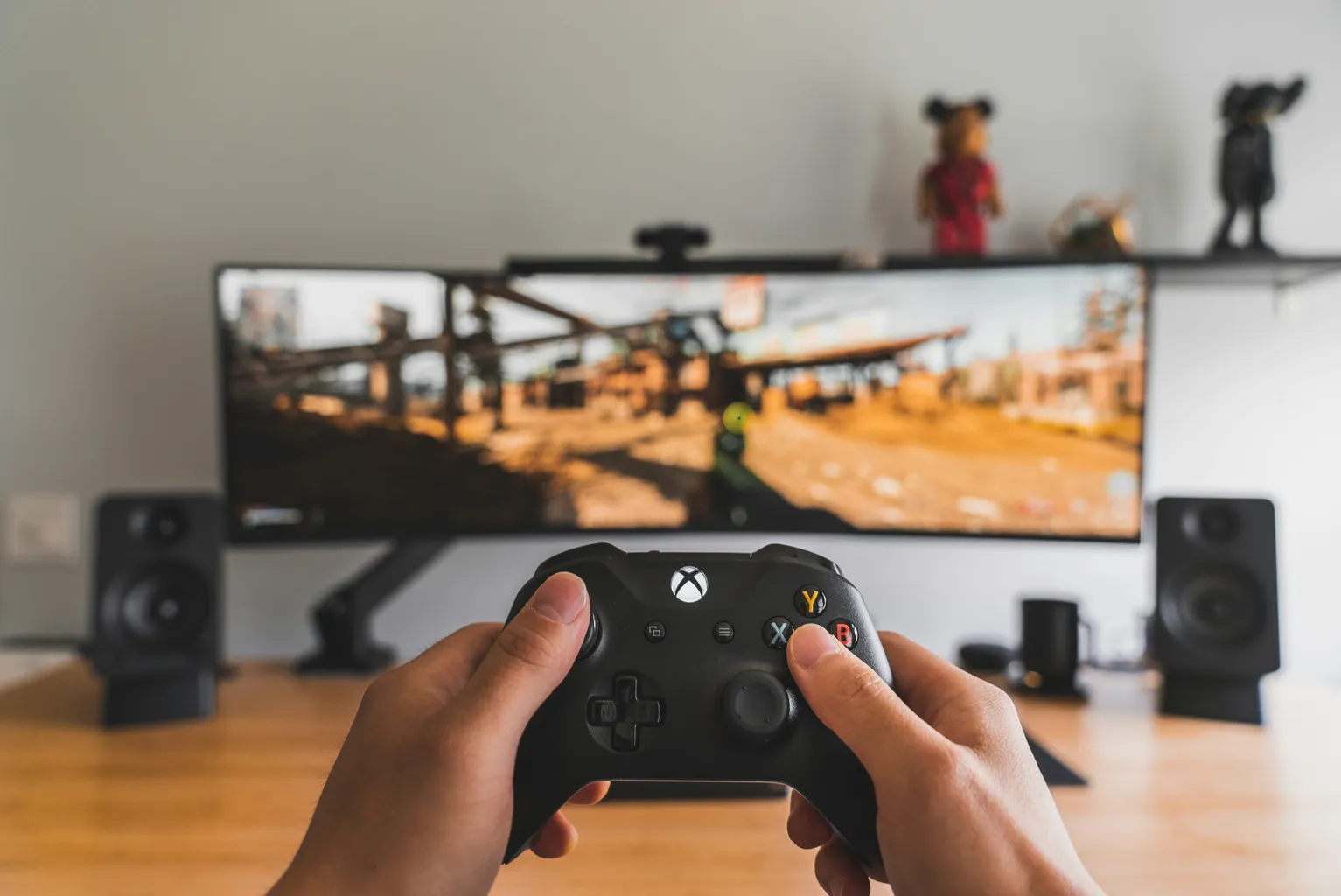 Person holding a black Xbox controller playing a video game on a widescreen monitor with speakers on a wooden desk.