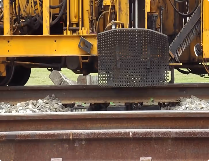 Close-up of a mechanical component with red springs, positioned above railroad track gravel and a wooden railroad tie.
