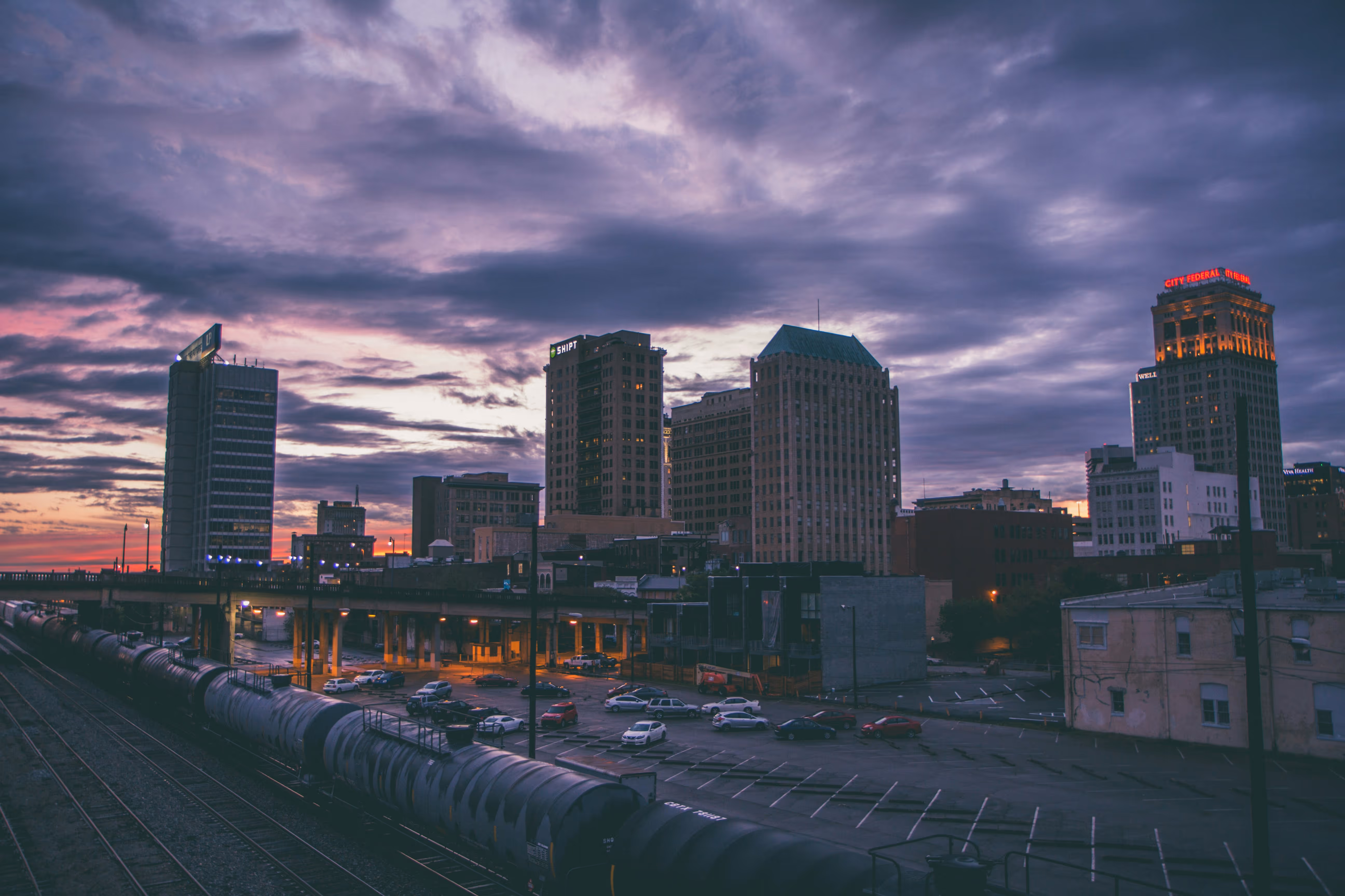 Urban skyline at dusk with dark clouds, illuminated buildings, parked cars, and a freight train on tracks in the foreground.