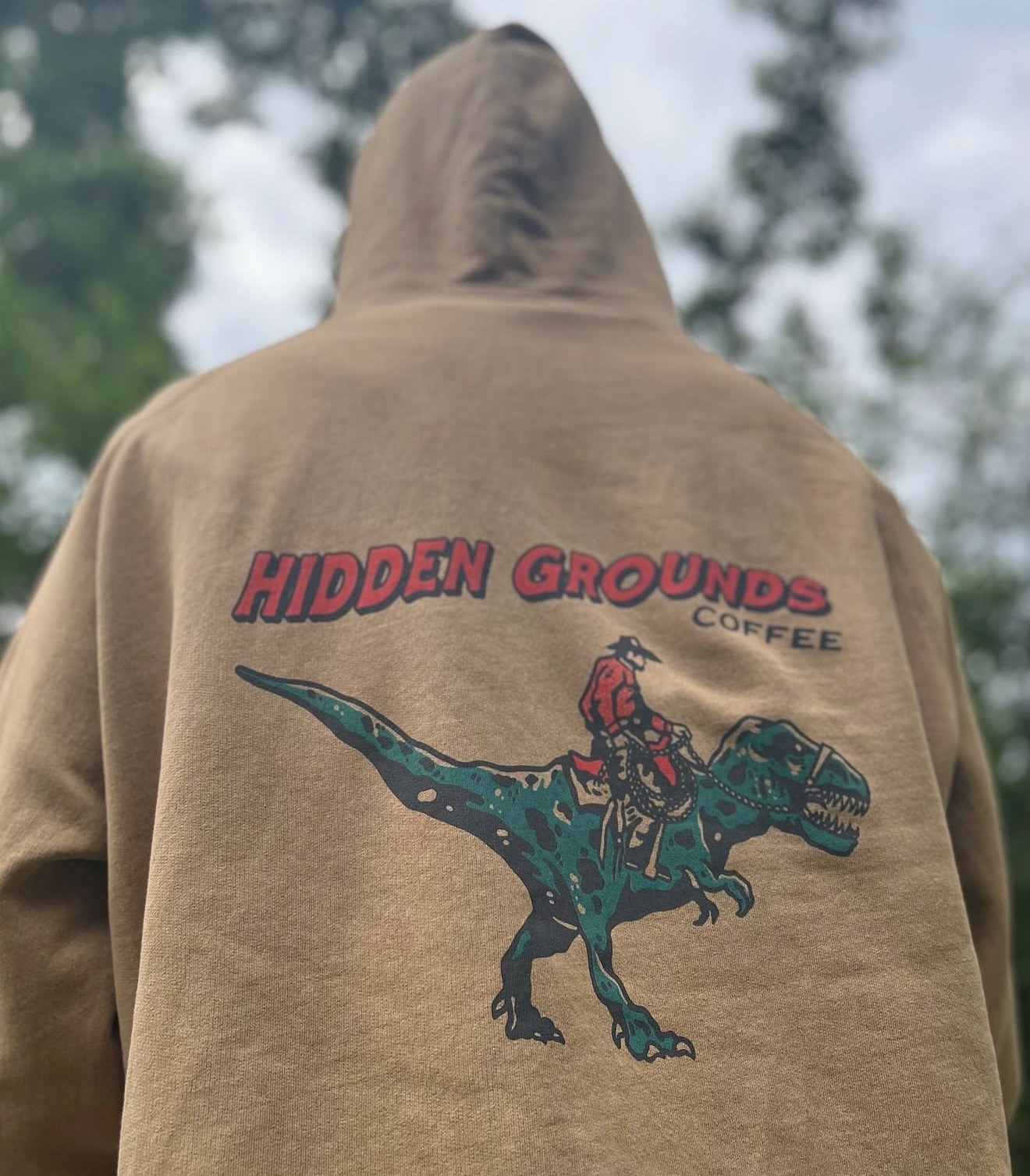 Brown hoodie with a graphic of a person riding a dinosaur and the text 'HIDDEN GROUNDS COFFEE' on the back.