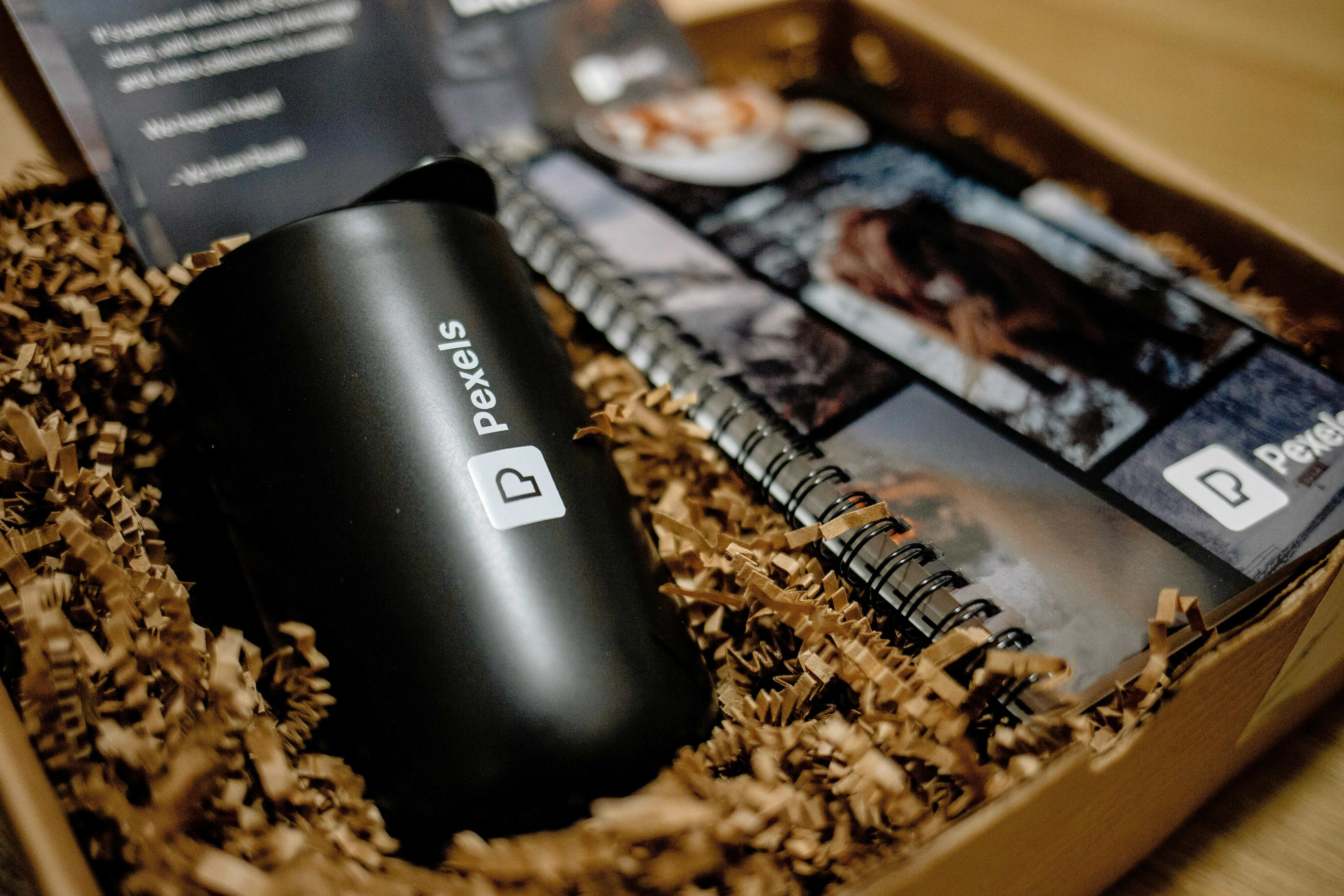 Black Pexels-branded tumbler and a spiral notebook with Pexels branding nestled in brown packing paper inside a box.