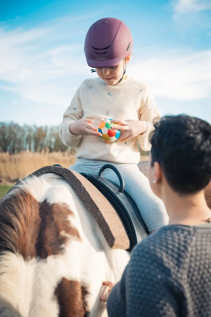 Ein Kind sitzt auf einem Pferd und spielt mit einem bunten Ball.