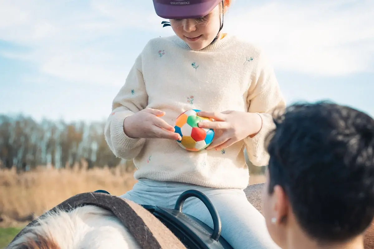 Ein Kind sitzt auf einem Pferd und spielt mit einem bunten Ball.