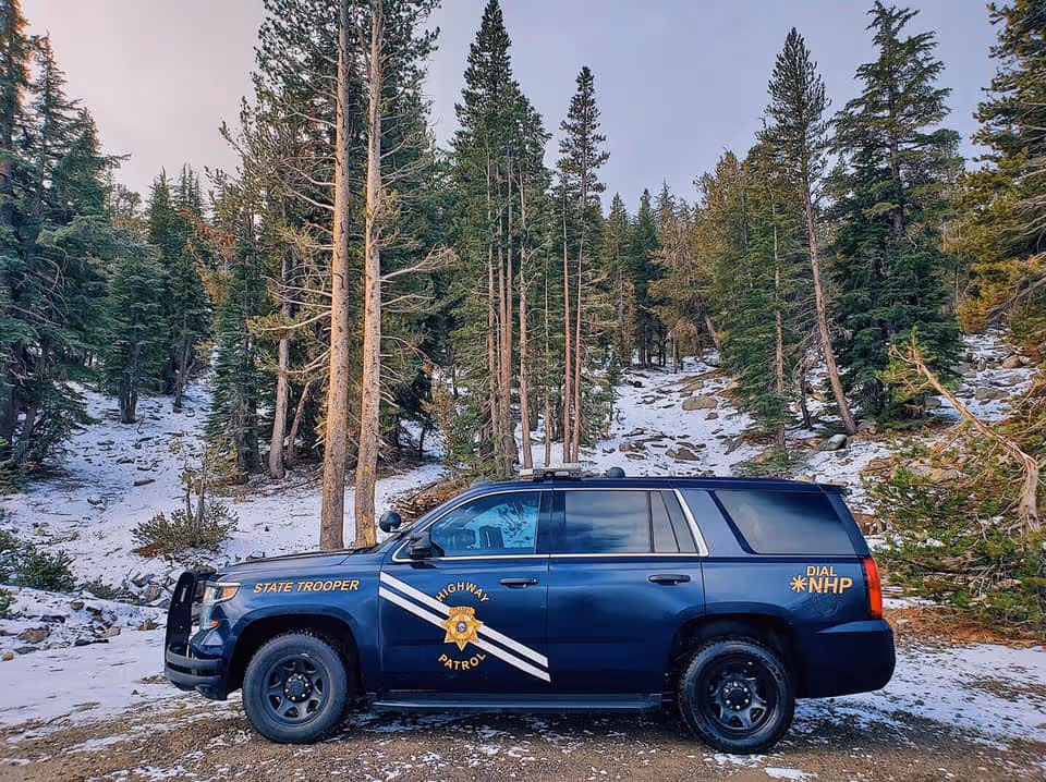 Blue New Hampshire State Trooper highway patrol SUV parked on snowy forest ground surrounded by pine trees.