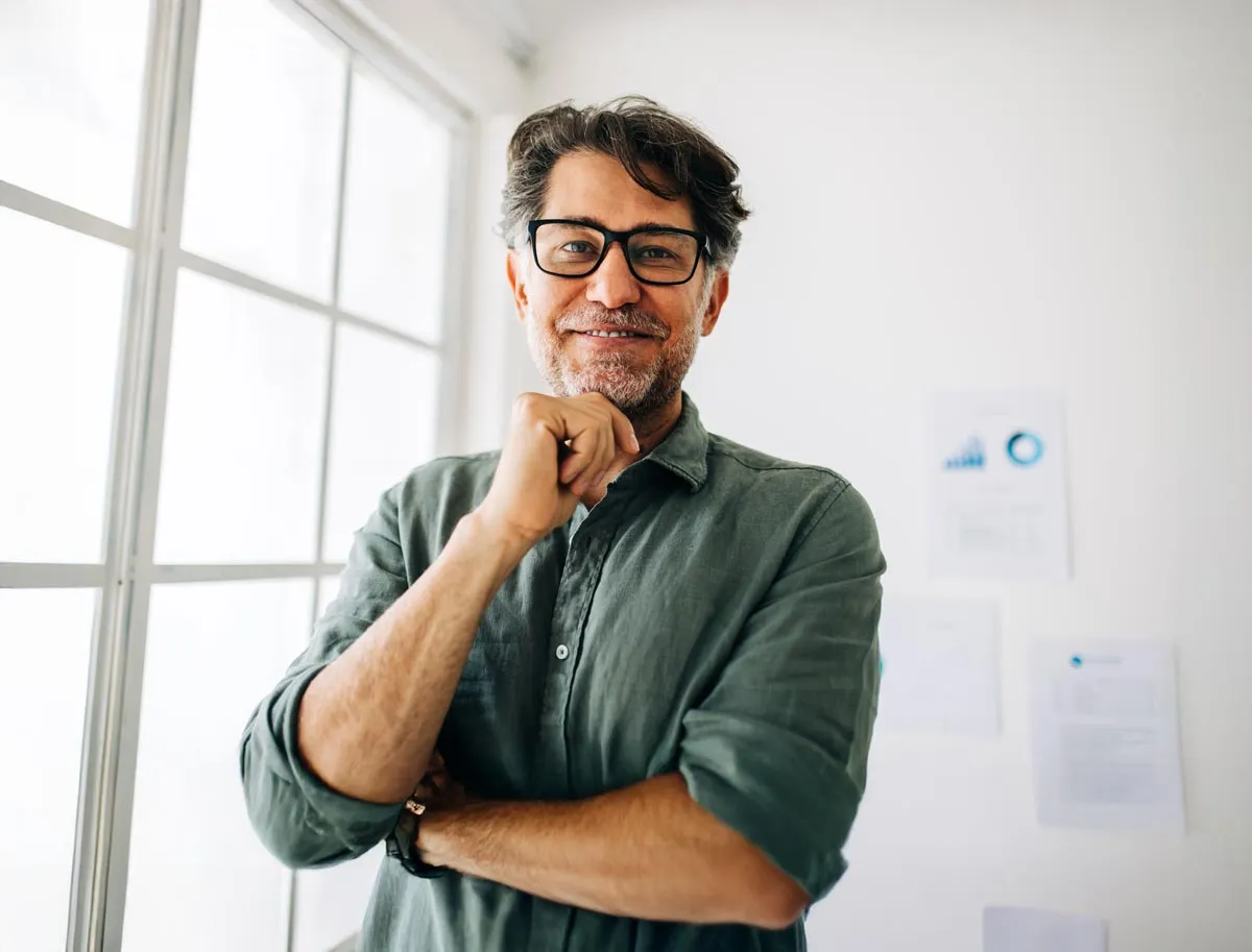 Smiling middle-aged man with glasses and gray beard standing with folded arms in a bright room.
