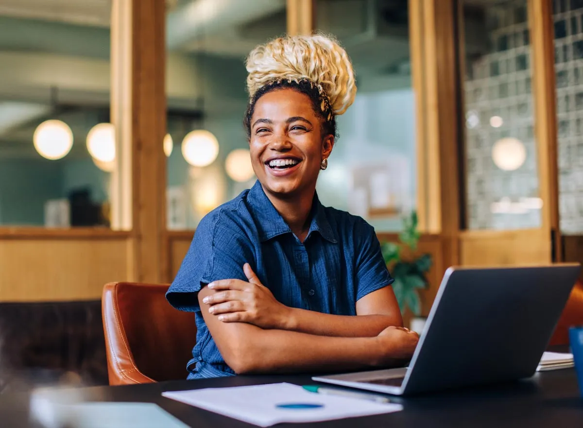 Smiling woman with blonde hair in a bun sitting at a desk with a laptop in a modern office.