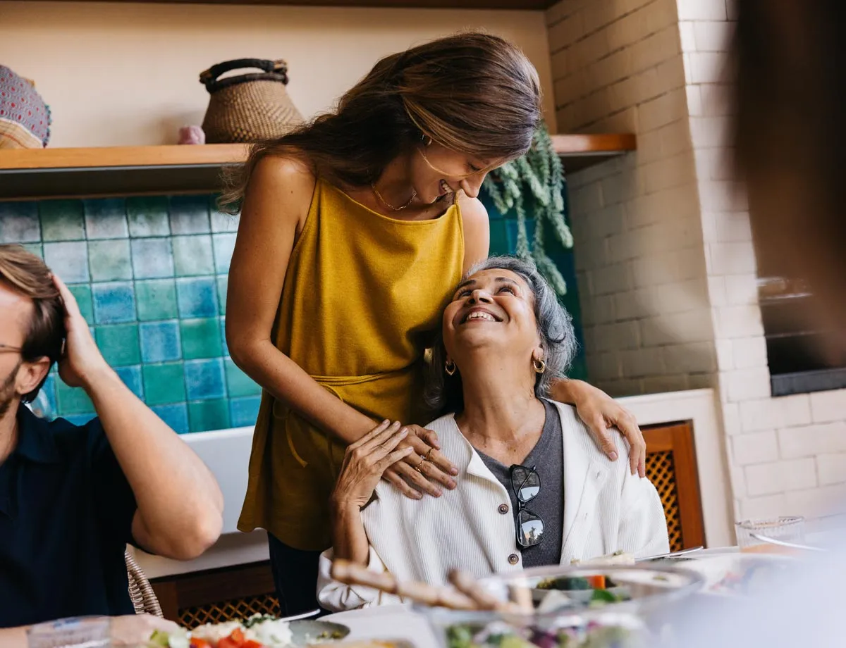 Younger woman in yellow top hugging and smiling at an older woman seated at a dining table.