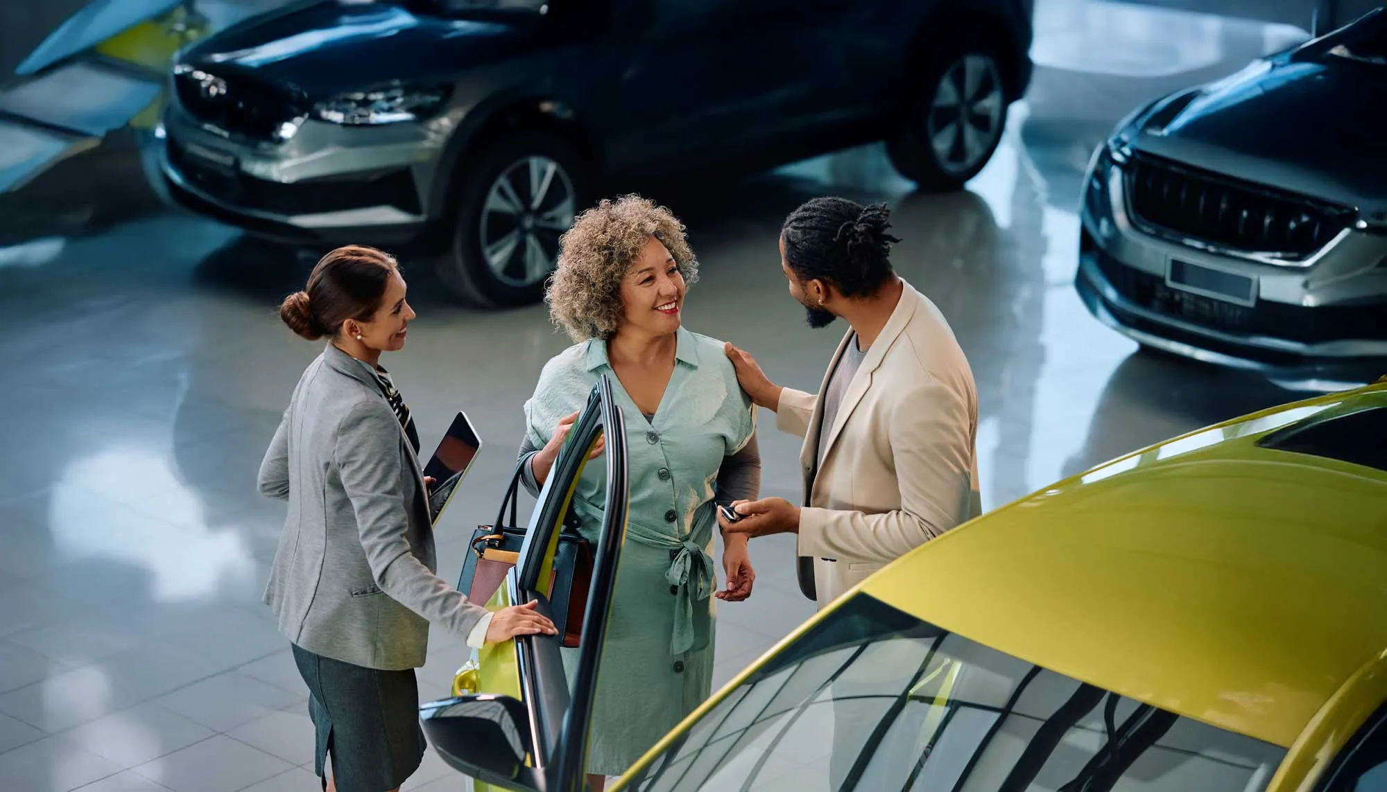 Two car salespeople assisting a smiling woman as she looks at a yellow car in a showroom.
