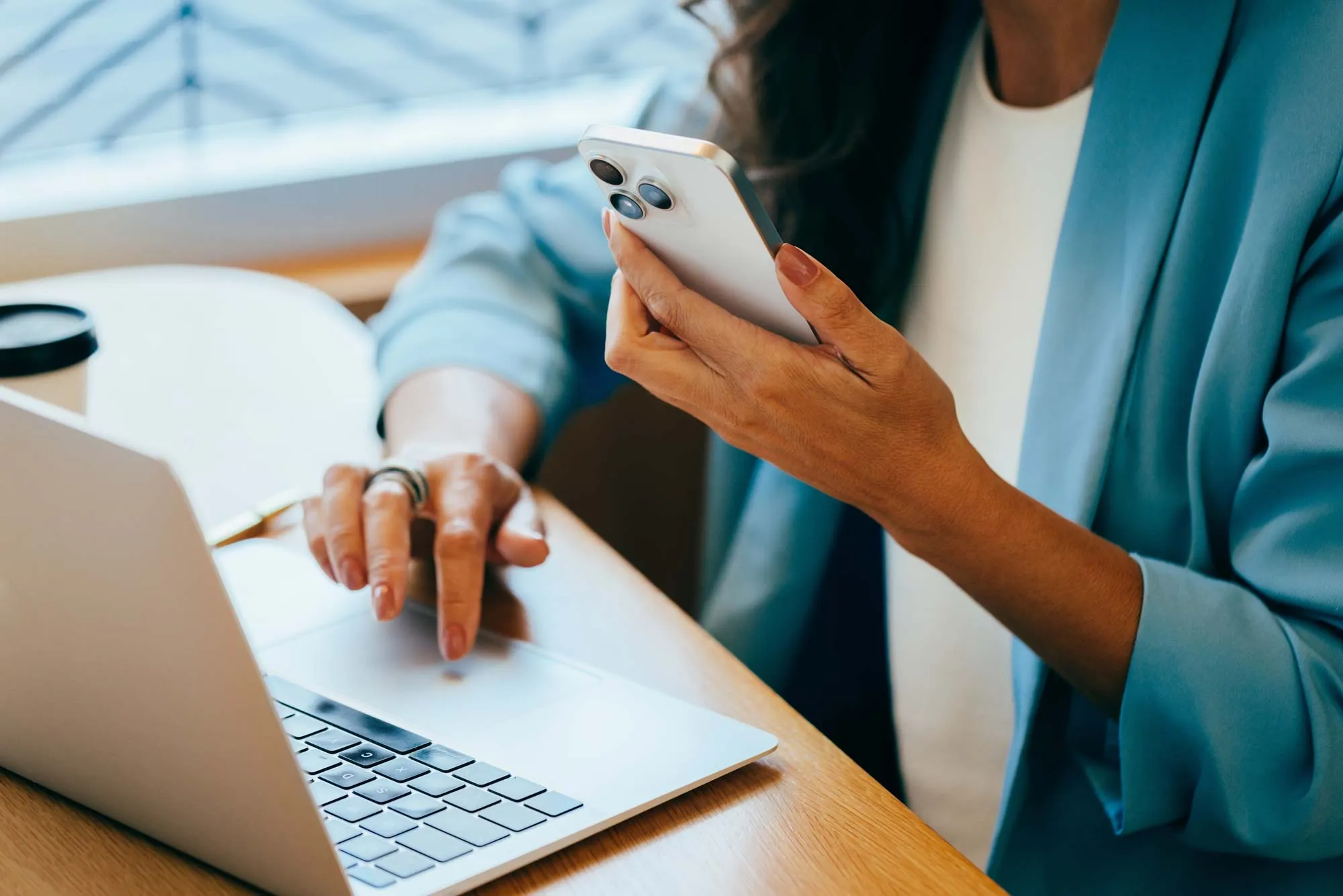 Person in blue blazer using smartphone with one hand and operating laptop touchpad with the other on a wooden desk.