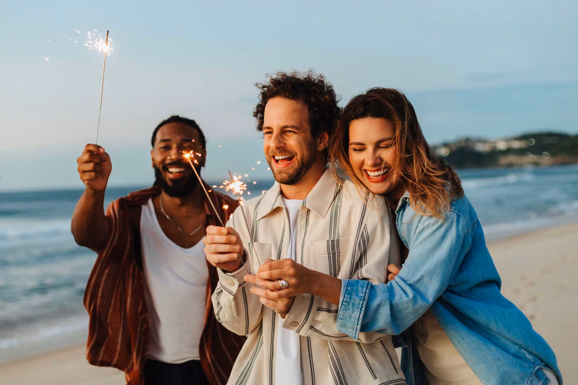 Three friends smiling and holding sparklers on a beach at sunset.