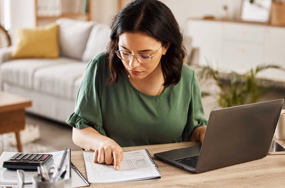 Woman wearing glasses and a green top working at a desk with a laptop, calculator, and documents.