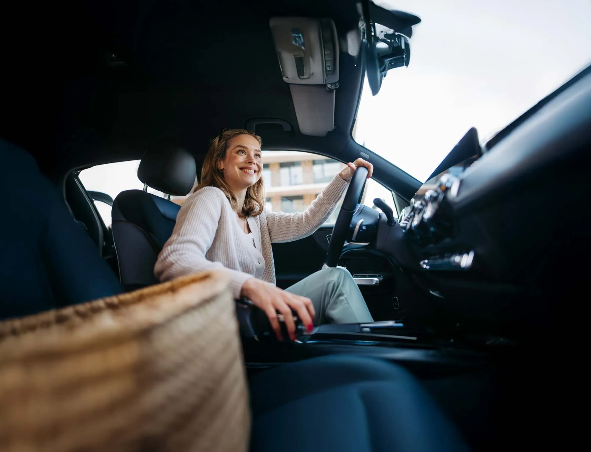 Smiling woman sitting in the driver's seat of a car, holding the steering wheel with one hand.