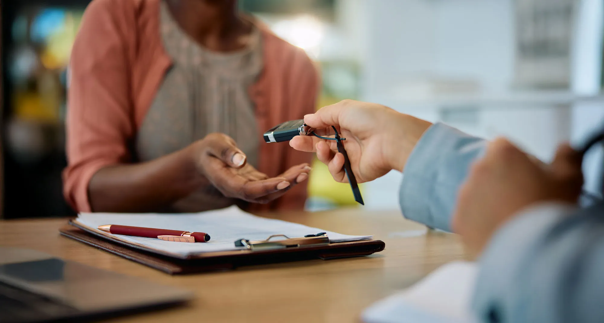 Person handing over car keys to another person sitting at a desk with a clipboard and pen.