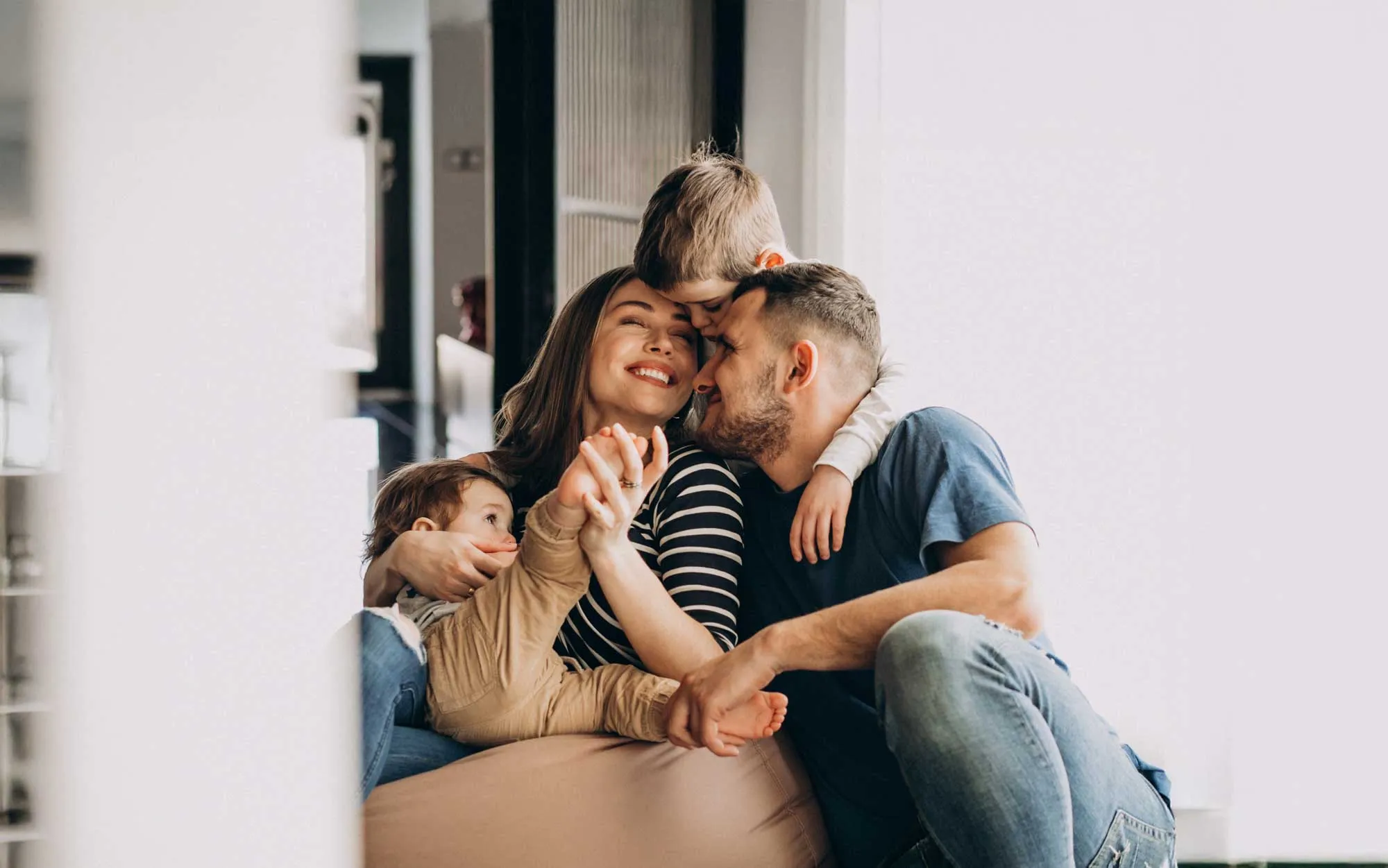 Smiling family of four cuddling closely together inside a home with natural light.