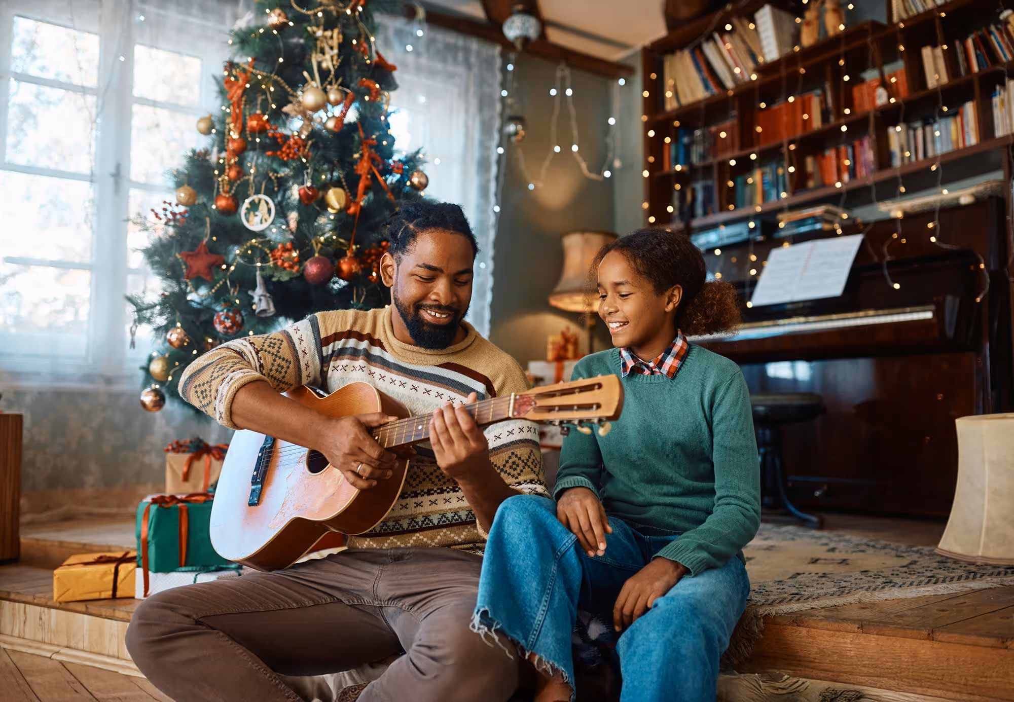 Man playing guitar while sitting next to smiling girl in a living room decorated with a Christmas tree and lights.