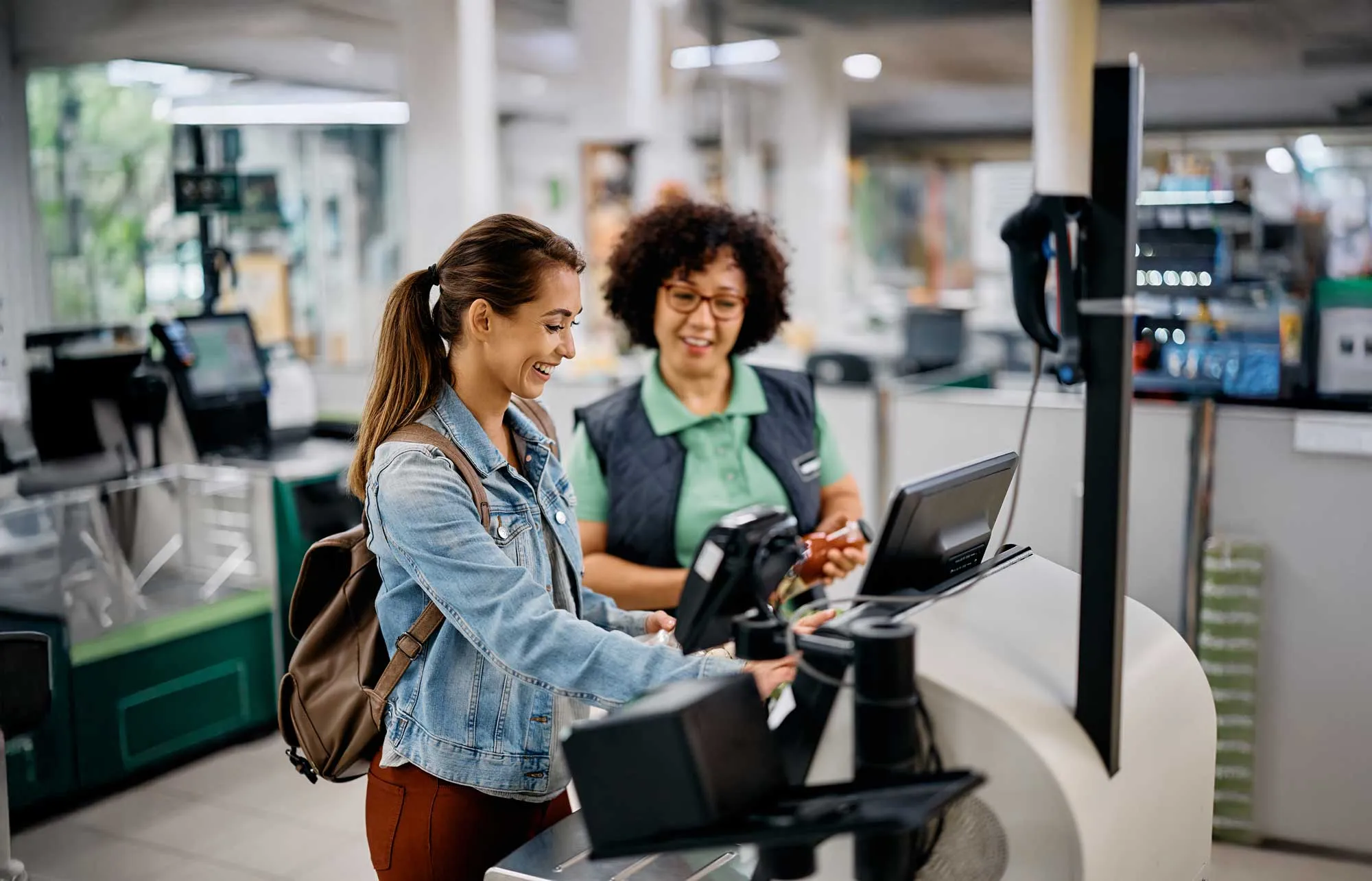 Smiling young woman using a card payment terminal at a grocery store checkout with a cashier in the background.
