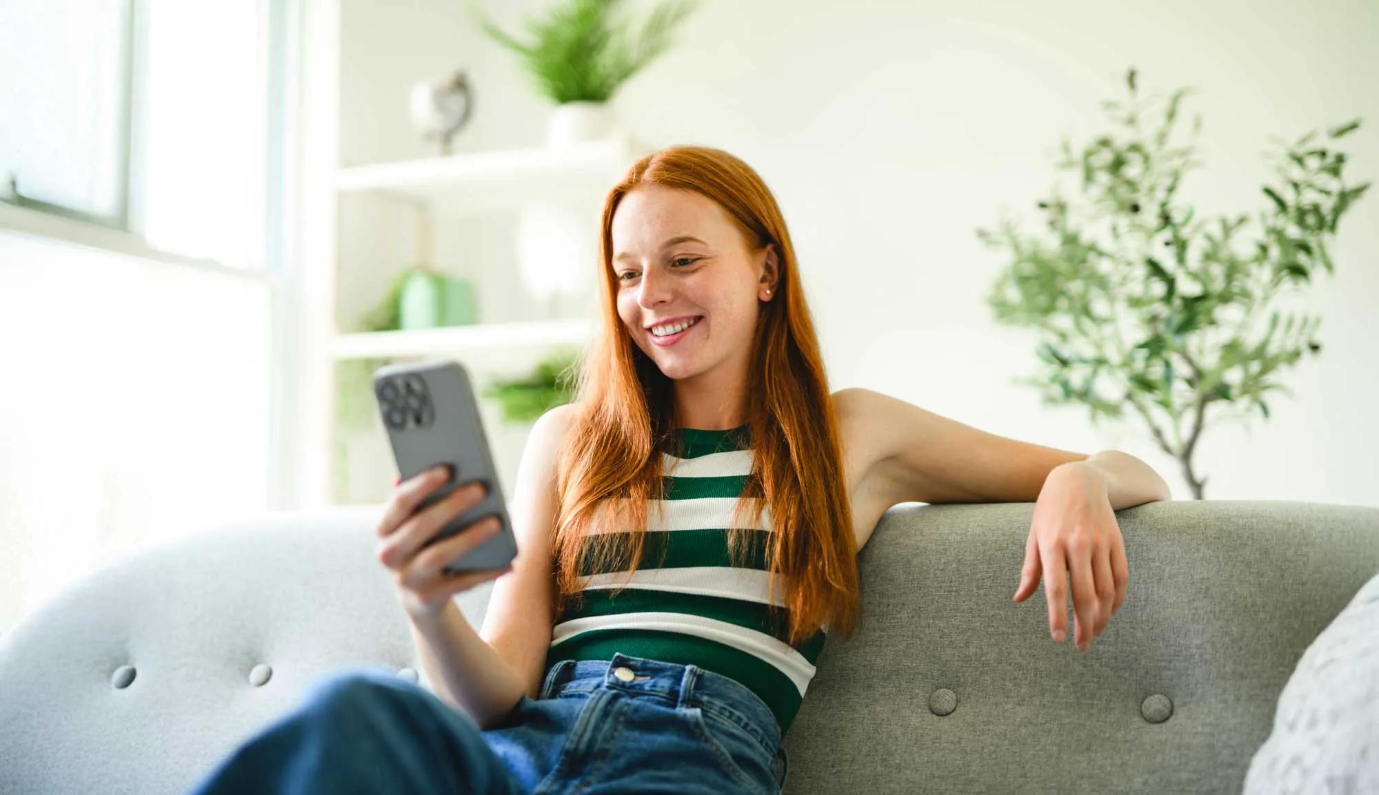 Young woman with long red hair sitting on a gray sofa, smiling and looking at her smartphone.