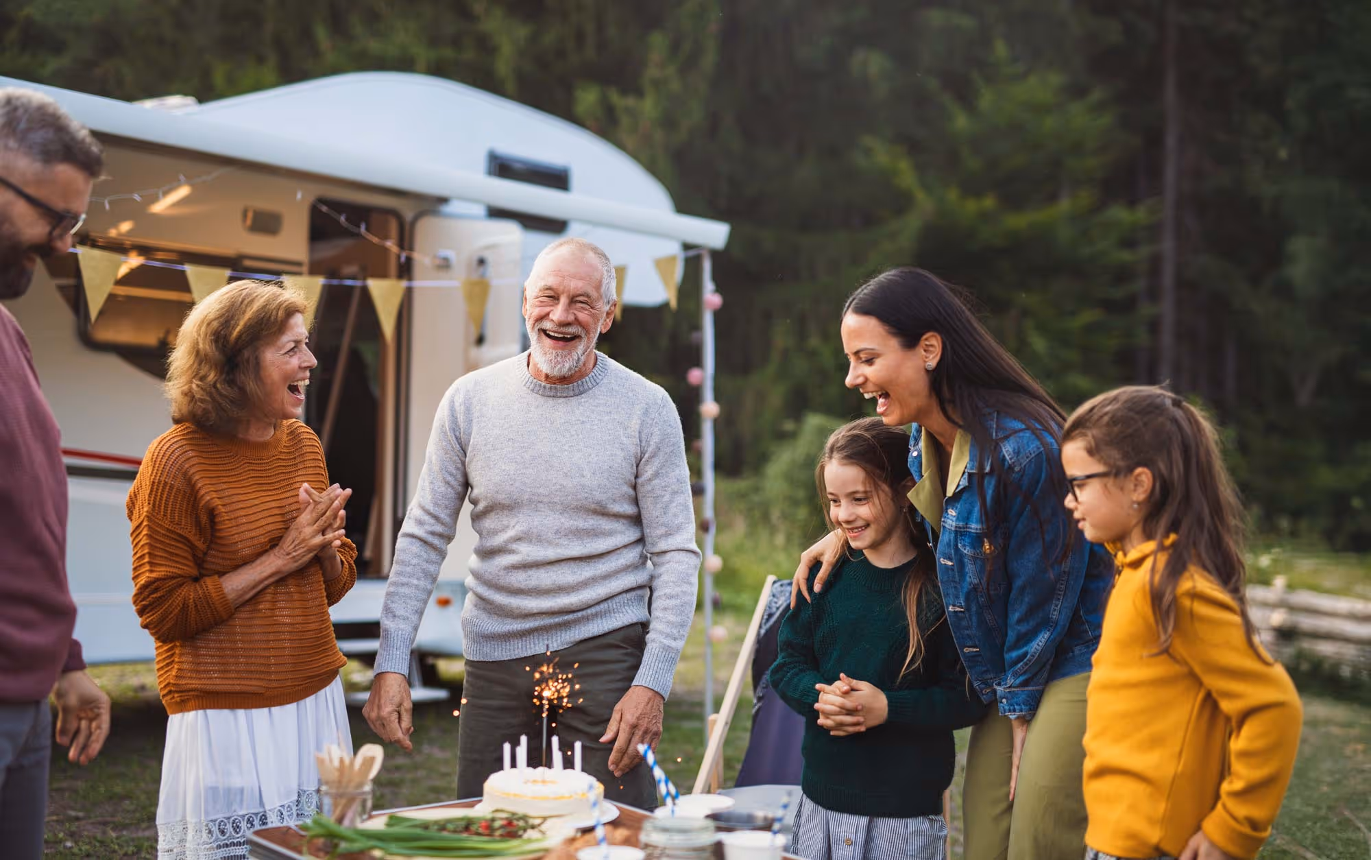 Family celebrating a birthday outdoors near an RV with a cake and sparklers on the table.