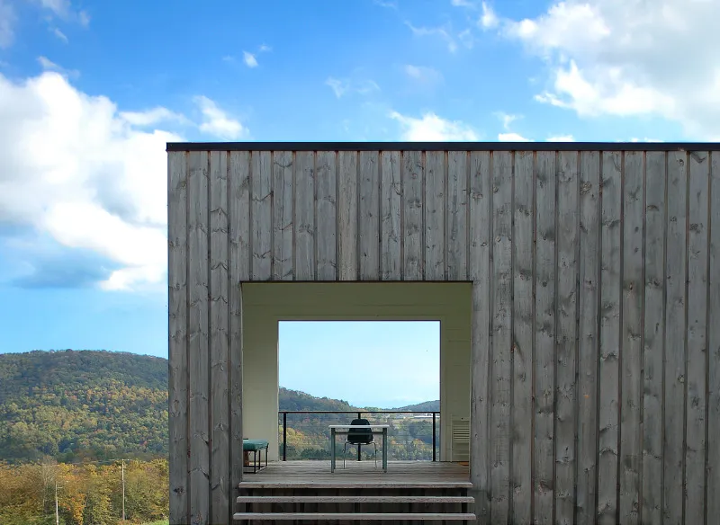 Modern wooden cabin with open rectangular doorway in  grassy field overlooking hills.