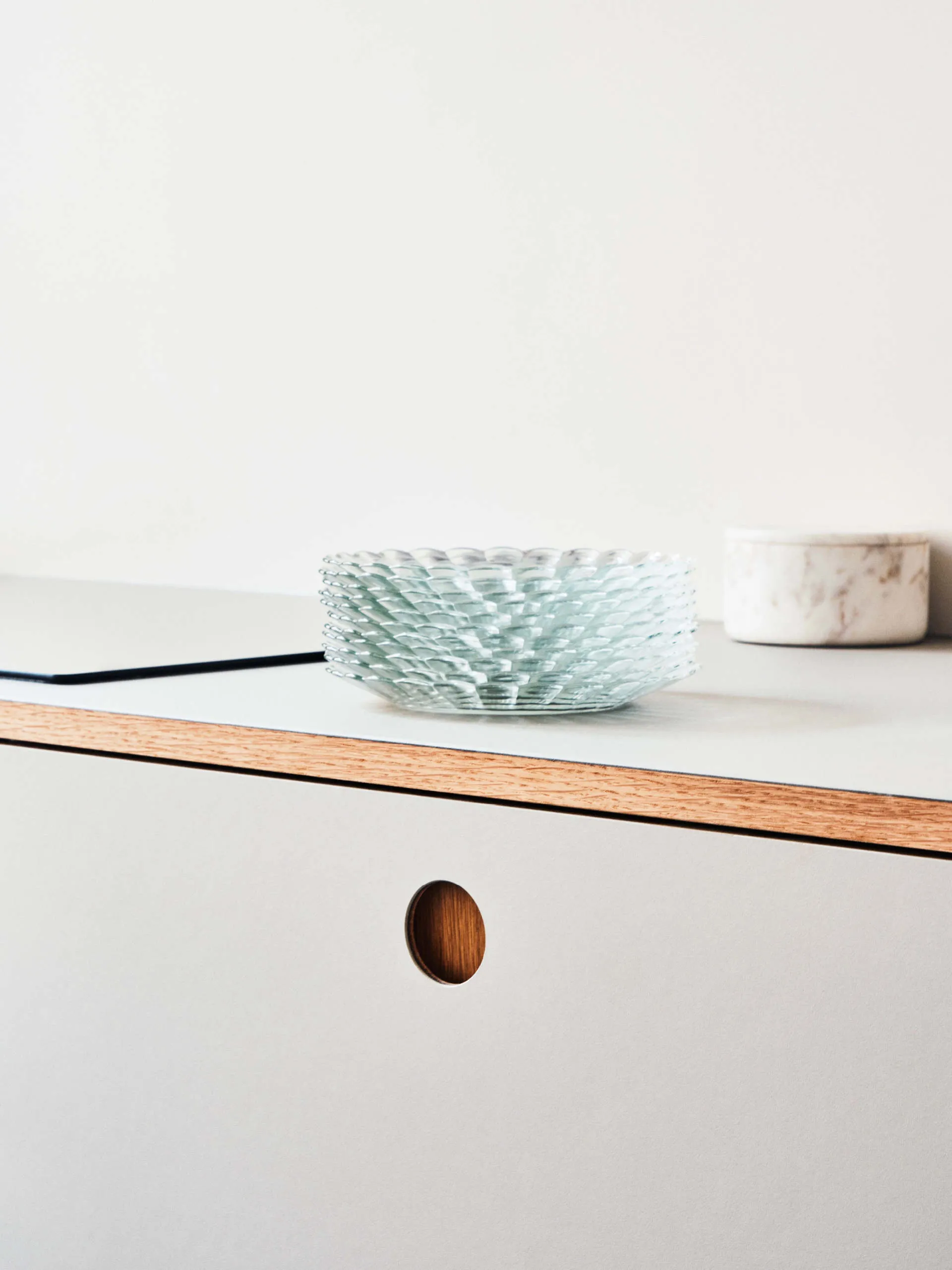 Minimalist countertop with a stack of clear textured glass plates, a marble container, and a white cabinet with a circular wooden cutout handle.