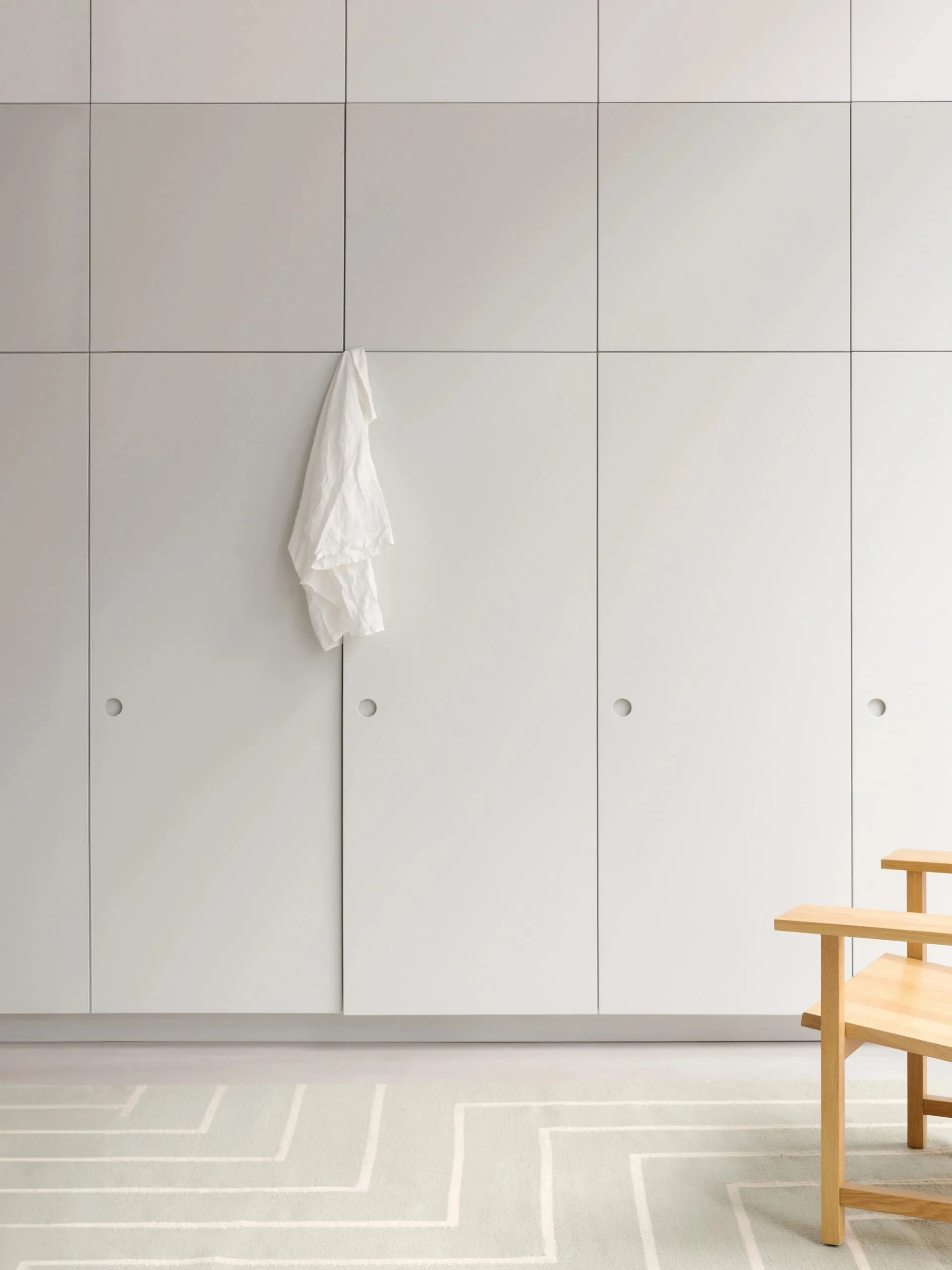 Minimalist white cabinetry with a white towel hanging on one door and a light wood chair on a rug.