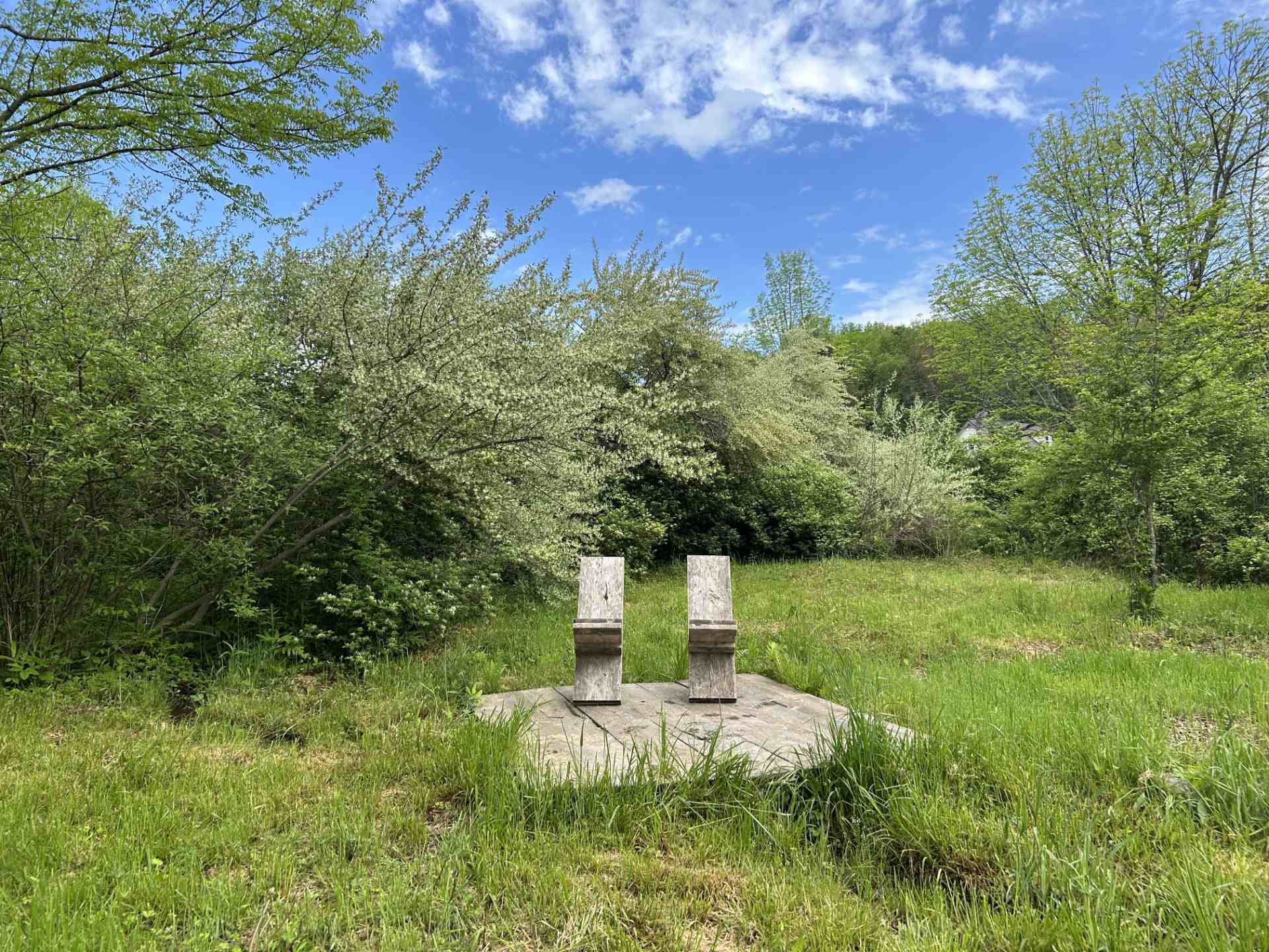 Two weathered wooden chairs on a raised wooden platform in a grassy clearing surrounded by trees.